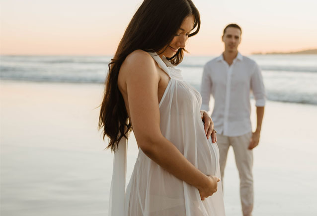 pregnant women at the beach holding her bump with husband looking on