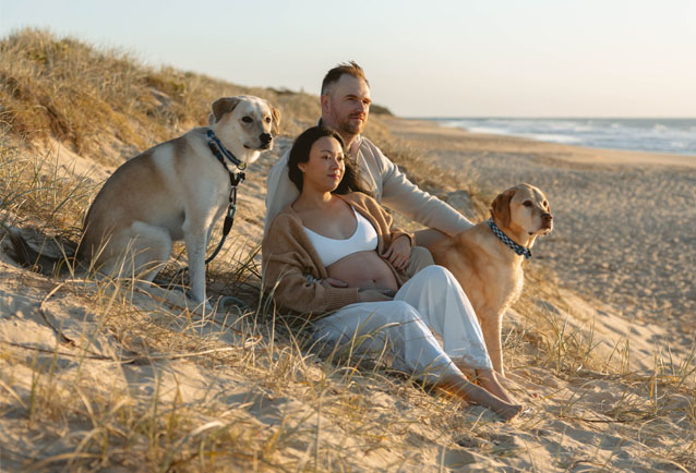 pregnant woman sitting down at the beach with her husband and dogs